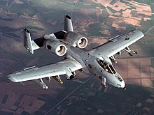 Aerial top view of gray jet aircraft flying above green and brown patchy earth surface. Under each wing are eleven hardpoints for various weapons and ordnance. The two engines are located aft of the wings and in front of two fin units.