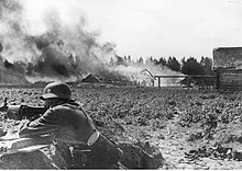 Soldat allemand allongé au sol avec une mitrailleuse devant des maisons en feu