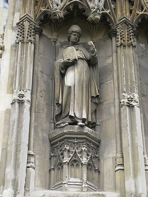 Stone statue of a standing man wearing robes and a mitre carrying a tablet in one hand and holding his other hand up.