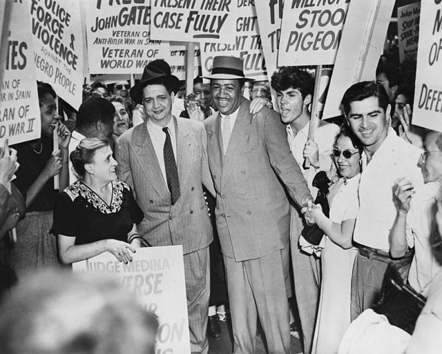 Two men dressed in suits are surrounded by people holding signs.