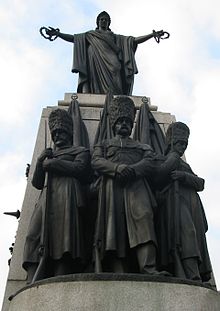 Photographie d'une statue en bronze d'une femme en toge tenant des couronnes de lauriers au sommet d'un pilier blanc. Au pied de cette dernière, trois statues représentent des soldats avec de longs uniformes et des bonnets noirs en poil d'ours.