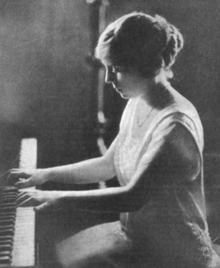 A young white woman, seated at a piano, hands on the keys; her hair is dressed to the back of her head; she is wearing a light-colored gown with bare forearms and a scooped neckline