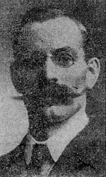 Black and white photo of a middle-aged man wearing eyeglasses, a handlebar moustache, white-collar dress shirt and a necktie