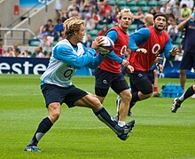 Un sportif avec un chasuble blanc observe le jeu avant de jouer le ballon qu'il tient des deux mains, des adversaires en chasuble rouge en défense se rapprochent.