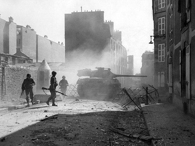 United States Army infantry supported by a M18 tank destroyer advancing through an enemy-occupied town during World War II, the most recent conflict t