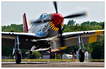 A single nosed-propeller aircraft is on the ground on its wheels with the propeller in motion. The aircraft is viewed from the front, but the red nose faces slightly to the right. The aircraft has black propellers. Parts of the wings and propellers are cropped from view.