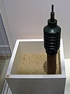 Schwarze, keulenförmige Landmine mit einem Dorn am oberen Ende, montiert am Ende eines Holzpfahls, der in einem mit Sand gefüllten Behälter steht.