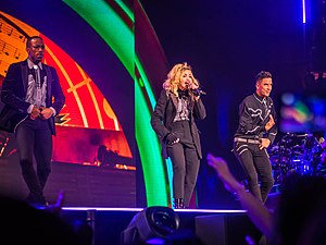 Madonna onstage wearing a black suit. She's flanked by two male dancers dressed in a similar style. The backdrop behind them shows orange and green flashes.