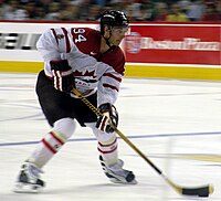 Hockeyspeler in het witte uniform van Canada.  Met zijn stok loodst hij een puck over het ijs.