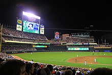 Terrain de baseball la nuit, le tableau de bord affiche un joueur, les jumeaux "M" et "Stp" décorent une grande enseigne au néon