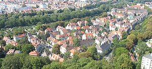 In the upper part of the picture from left to right above (north) the Wertach, lined on both sides by tall trees with Pfersee above in the left half of the picture, almost to the right the Rosenaustrasse a little diagonally also under a row of trees and a smaller one in front of the multi-storey houses on the roadside, in between the city district from above with different shades of red and roofs.  Some trees between the houses, the green city park below the hotel tower in the photo below