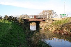 A narrow drain crossed by a small bridge. The bridge is of rusted, and untreated steel, and consists of a straight steel beam supporting the road deck, with, behind it, a steel arch. The arch is fixed in a buttress of dressed and coursed stonework, visible only on the far bank. There is another on our side of the drain, but it is obscured by vegetation. Rusty handrails protect pedestrians from falling. The water is flat, reflecting (where it is not in shadow), the blue sky above.