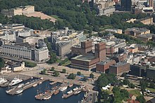 Luftbild eines an der Küste gelegenen Stadtviertels. Im Meer liegen Boote, an Land ist ein Platz, dahinter mehrere größere Gebäude