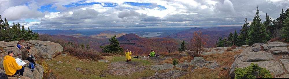 A wide open ledge, with small coniferous trees on either side, on which several people are sitting down and reading, eating or taking photographs. Beyond it lies a mountainous region, with many peaks in fall color. A large body of water is visible just right of center, at the foot of some smaller mountains.