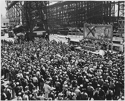 The AJC Band, from Hamilton Field, plays at a war bond rally held at Mare Island on 26 June 1945. Behind the band, caricatures of Benito Mussolini and Adolf Hitler have been crossed out and a fanged Japanese figure is labeled "Tough One To Go"