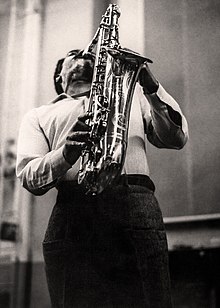 Photograph of a man with a short afro hairstyle and short goatee playing saxophone, arching his back to raise the instrument above his head