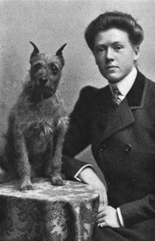 A young white woman in a tailored suit and necktie, sitting at a small table next to a wire-haired dog.