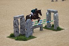 Un cavalier avec une veste verte et un pantalon blanc montant un cheval bai franchit un obstacle représentant un mégalithe surmonté de barres.