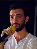 A 35-year-old man with dark hair and a dark beard talks into a microphone, looking towards the left of the camera.