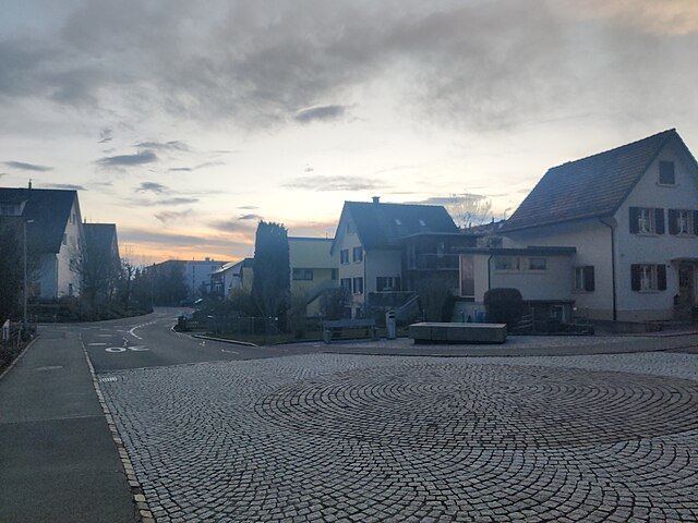 The concrete fountain which today is located at the intersection of Zihlstrasse and Gstaldenstrasse in Hinwil was designed by Max Vogt as part of the Hinwil railway station. It has since been moved away from the railway station and placed here. The photo shows the fountain from the northeastern side of the intersection, overlooking both roads and facing west. The residential building to the right is Gstaldenstrasse 20