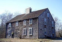 A traditional 2-story colonial-style house, with weathered cedar shake siding and roofing. A woman stands near the entrance.