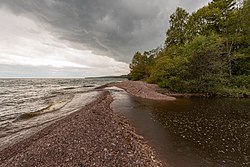 Ein Strom dunklen Wassers fließt von der rechten Seite der Bilder weg von der Kamera und einige Wege einen steinigen rötlichen Strand hinunter, vorbei an einem Wäldchen grüner Bäume, bevor er links über den Strand biegt und auf einen See trifft, der so breit ist, dass er weit entfernt ist kann nicht gesehen werden. Der Himmel ist auf der rechten Seite wolkiger, weniger links über dem See.