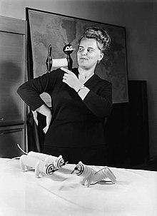 black-and-white image of Libuše Niklová with one of her toy cats perched on right shoulder, three more toy cats sitting on table in front of her