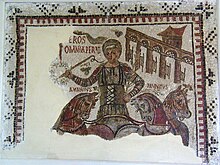 Mosaïque représentant, de face, un aurige sur son char, tenant une couronne. Trois chevaux sont visibles, dont deux surmontés de leur nom. En haut, à droite, les portes d'entrée de la piste