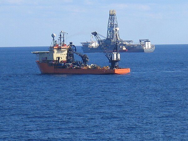 The drillship Discoverer Enterprise is shown in the background, at work during exploratory phase of a new offshore field. The Offshore Support Vessel 