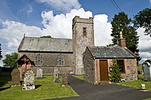 Foto av en kirke med et lite tårn festet til en lavere bygning.  Til venstre er det gravsteiner, mens det til høyre er en liten bygning foran kirken med en brun dør.