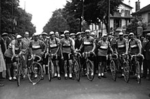 Photographie en noir et blanc d'une équipe cycliste au départ d'une course, les coureurs se tenant debout à côté de leur vélo.