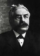 Tre-kvart længde fotografisk portræt af en mand med gråhvidt hår, buskede øjenbryn og overskæg, iført briller, slips, hvid skjorte, blazer og sort jakke