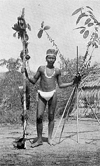 Fotografía en blanco y negro de un hombre con taparrabos y plumas con un palo en una mano y cuatro palos largos en la otra.