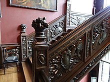 A photograph of the dark wooden panelling of the staircase, engraved with military symbols, and the carved baskets of fruit which sit atop the newel post at the bottom of the baluster