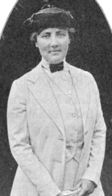 A white woman, standing, wearing a hat and a light-colored three-piece tailored suit