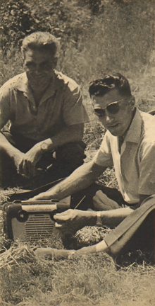 Deux hommes assis, en bordure d'un champ, avec un poste radio à leurs pieds