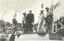 Cinco personas en la plataforma de piedra, de izquierda a derecha: hombre con bigote con uniforme militar y sombrero negro alto y tupido con barbijo;  hombre con uniforme militar con muchas medallas y sombrero de oficial naval anticuado;  hombre de pelo blanco con traje con abrigo largo oscuro y cuello blanco;  mujer vestida de estilo victoriano con sombrero blanco;  hombre con barba y pelo salvaje en traje.  Multitud y banderas en segundo plano.