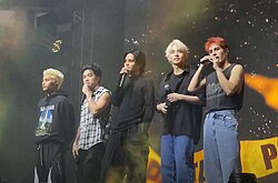 A group photo of SB19 on stage, each holding a handheld microphone.