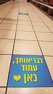 Tiles on supermarket aisle floor. Every seven tiles there is a sticker saying "For the sake of your health, stand here" in Hebrew.