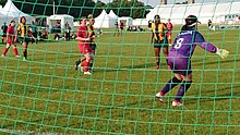 Durch ein Fußballtor sieht man die Torhüterin der kanadischen Mannschaft im lila Dress von hinten, auf dem Spielfeld mehrere Spielerinnen der kanadischen Mannschaft im roten Trickot und der südafrikanischen Mannschaft in Grün-Gelb. Im Hintergrund, am Rand des Spielfelds, sind weiße Zelte aufgebaut.