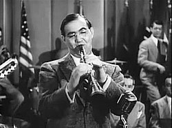 A Caucasian man in his thirties is standing and playing the clarinet, facing the camera. His dark hair is parted to the side and he is wearing glasses. Both of his hands are on the clarinet and he is blowing into the instrument with his eyes partly closed. There is a microphone on the foreground next to the bell of the clarinet. Several other musicians can be partly seen on the background. In the corner on the right hangs a flag with white and red stripes and white stars on a blue background.