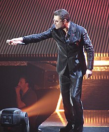 Photographie couleur d'un homme sur une scène de concert qui tend un micro au public.