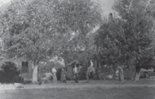 una foto en blanco y negro de una granja en Utah en la década de 1870.  La casa está cubierta por algunos árboles y cuatro personas se paran frente a ella.