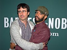 Photo of Oliver, wearing a grey hoodie, embracing Cenac, who is wearing a red hoodie. Both are standing in front of a press backdrop labeled "Barnes & Noble".