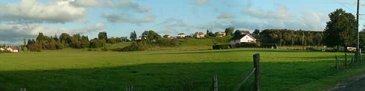 Un champ derrière lequel s'élève une petite collines plate ponctuée de maisons et d'arbres.
