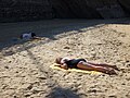 Two women, who are not together, sleeping on towels on sand at a beach, in 2015. The women’s bare feet hang over the ends of their towels. One of their faces is clearly visible.