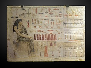 A flat limestone block with a painted, carved raised-relief of woman in spotted linen cloth, seated near table with food items. Painted hieroglyphs decorate the rest of the surface.