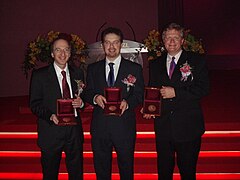 Zdjęcie trzech mężczyzn, ubranych w stroje wizytowe, trzymających przed sobą wyścielane czerwonym aksamitem pudełka z medalami. Zdjęcie wykonano podczas uroczystości, w tle widać podświetlone czerwone schody oraz bukiety kwiatów.