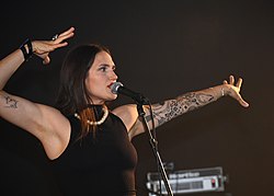 Marina Maximilian Blumin wearing a black top, standing in front of and appearing to speak or sing into a microphone, gesturing with both arms