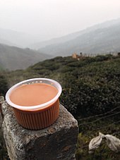 Vordergrund: Plastikbecher mit Darjeeling-Tee mit Milch.  Zweiter Plan: Teeplantagen in Darjeeling.